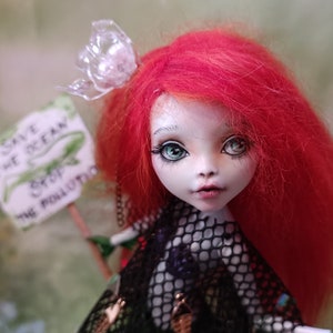 May include: A handmade doll with bright red hair and green eyes. The doll is holding a sign that reads "Save the Ocean, Stop the Pollution." The doll is wearing a black mesh top and a clear plastic butterfly hair accessory.