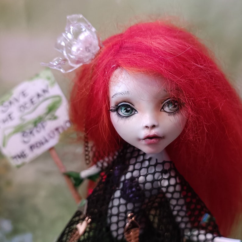 May include: A handmade doll with bright red hair and green eyes. The doll is holding a sign that reads "Save the Ocean, Stop the Pollution." The doll is wearing a black mesh top and a clear plastic butterfly hair accessory.