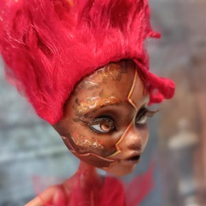May include: A custom doll with a fiery red wig and detailed face paint. The doll's skin tone is dark, with intricate patterns in shades of brown, gold, and orange. The doll has large, expressive eyes and long eyelashes, with a red mesh garment.