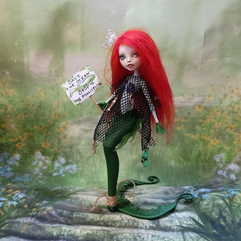 May include: A handmade doll with red hair and green clothing, holding a sign that reads "Save the Ocean Stop the Pollution." The doll has a fish tail and is wearing a black net over its shoulders. The background is a blurred image of a green field.