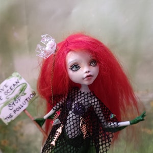 May include: A custom doll with red hair, green skin, and a black net top. The doll holds a sign that reads "Save the Ocean, Stop the Pollution." Gold fish-shaped charms adorn the doll's outfit. A unique art piece.