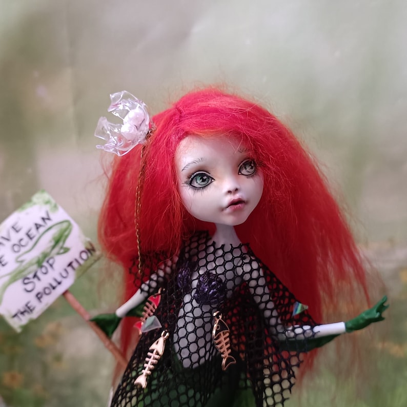 May include: A custom doll with red hair, green skin, and a black net top. The doll holds a sign that reads "Save the Ocean, Stop the Pollution." Gold fish-shaped charms adorn the doll's outfit. A unique art piece.