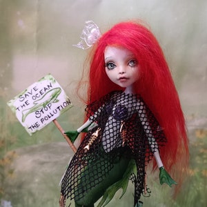 May include: A handmade doll with red hair and green eyes, dressed as a mermaid. The doll holds a sign that reads "Save the Ocean, Stop the Pollution." The mermaid wears a black net top and a green tail. A clear plastic flower adorns her head.