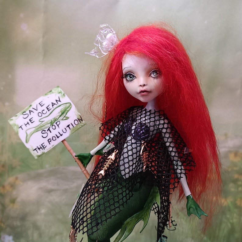 May include: A handmade doll with red hair and green eyes, dressed as a mermaid. The doll holds a sign that reads "Save the Ocean, Stop the Pollution." The mermaid wears a black net top and a green tail. A clear plastic flower adorns her head.