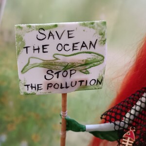 May include: A white sign with green border and text that reads "SAVE THE OCEAN STOP THE POLLUTION" with a green whale illustration. The sign is held by a green hand. The background is blurred.