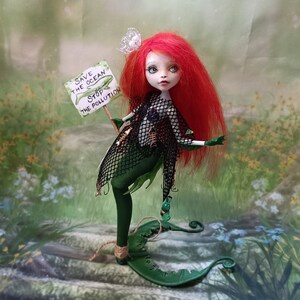May include: A handmade doll with red hair and green skin, holding a sign that reads "Save the Ocean, Stop the Pollution." The doll is wearing a black net top and a green skirt, with a green tail. The doll has a white flower in its hair.