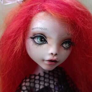 May include: A close-up of a doll's face with bright red hair and striking green eyes. The doll has pale skin, delicate features, and long, black eyelashes. The doll is wearing a black mesh top.