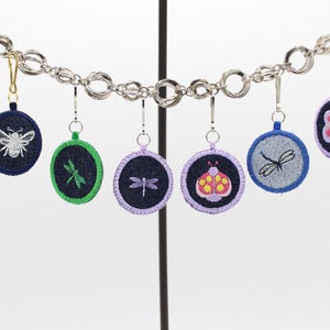 Puede incluir: Una colección de colgantes bordados con temática de insectos, colgados de una cadena plateada. Cada colgante presenta un diseño de insecto diferente, incluyendo una abeja, una libélula y una mariquita, con acentos coloridos. Los colgantes son redondos y tienen un fondo de mezclilla.