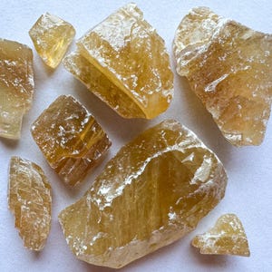 Puede incluir: Una colección de ocho cristales de calcita brutos, naturales, de color marrón dorado. Los cristales son de varios tamaños y formas, algunos tienen una textura estriada.