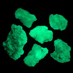 May include: Six green glow-in-the-dark rocks of various shapes and sizes. The rocks are arranged on a black background.