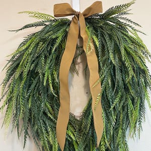 May include: A green and brown wreath with long, slender leaves. A brown ribbon is tied at the top, forming a bow and long hanging streamers. The wreath is hanging on a white surface.