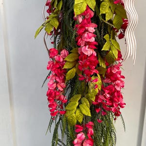 May include: A cascading floral arrangement featuring vibrant red and pink flowers, complemented by green leaves and long, slender green foliage. A striped ribbon adds a decorative touch. This artificial floral piece is suitable for home decor.