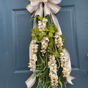 May include: A white and brown striped ribbon bow hangs from a door with a long, green garland of white flowers and leaves.