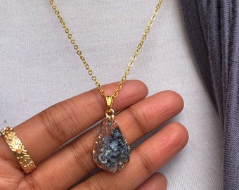 May include: A gold chain necklace with a teardrop-shaped pendant featuring a blue and gray crystal.