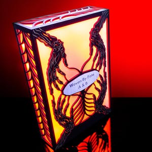 Op de afbeelding: Een rechthoekig, verlicht kunstwerk met een warmgele binnenkant en ingewikkelde, donkerrode en zwarte ontwerpen. De tekst "Woven By Fate A & B" staat op een ovale plaat. De achtergrond is een verloop van rood en zwart.