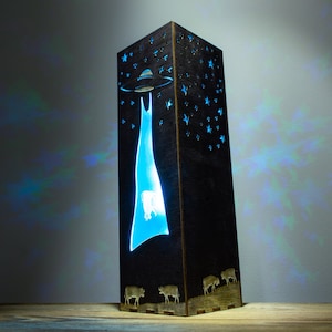 May include: A wooden lamp with a blue light shining through a cutout design of a UFO abducting a cow. The lamp is decorated with stars and has a dark wood finish.