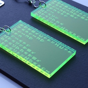 Könnte beinhalten: Ein Paar neongrüne, rechteckige Ohrringe mit eingravierten japanischen Schriftzeichen. Die Ohrringe hängen an silberfarbenen Haken und werden auf einer dunkelblauen Oberfläche präsentiert. Ein kleines weißes Etikett mit "Fawn Crafts" ist ebenfalls sichtbar.