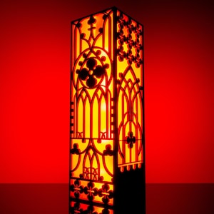 Op de afbeelding: Een hoge, rechthoekige lamp met een zwart, lasergesneden frame met ingewikkelde, herhalende geometrische en boogvormige patronen. Het interieur straalt een warme, heldere oranje gloed uit die de gedetailleerde uitsparingen verlicht.