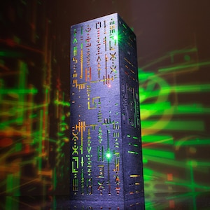 May include: A tall, rectangular, dark gray pillar featuring intricate, laser-cut geometric patterns and symbols. Bright green light shines through the cutouts, casting vibrant green and orange light streaks onto the surrounding area.
