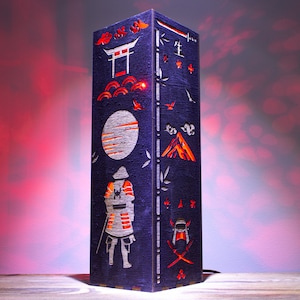 May include: A dark blue wooden lamp with a Japanese-inspired design. The lamp features a cut-out silhouette of a samurai warrior, a moon, a mountain, and a torii gate. The lamp is illuminated from the inside, casting a warm glow.