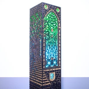 Op de afbeelding: Een hoge, rechthoekige, donkergekleurde lamp met gedetailleerde lasergesneden patronen. Het voorpaneel toont een oplichtend groen ontwerp in glas-in-loodstijl met een boom en een figuur, en een klein schildje met een blauwe leeuw.