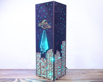 May include: A wooden lamp shaped like a city skyline with a UFO flying over it. The lamp is lit with a blue light, creating a futuristic and whimsical effect.