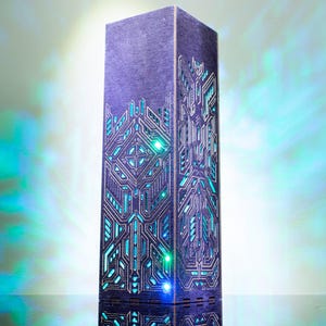 Peut inclure: Une lampe tour rectangulaire et haute en bois violet foncé, dotée de motifs géométriques complexes découpés au laser sur ses côtés. Les découpes révèlent une lumière interne brillante cyan et verte qui projette des motifs colorés alentour.