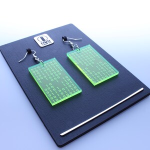 Pu&ograve; includere: Un paio di orecchini rettangolari verde neon con motivi a codice binario, esposti su una superficie nera. Gli orecchini hanno ganci argentati e sono su uno sfondo azzurro. Un logo argentato &egrave; visibile in alto a sinistra.
