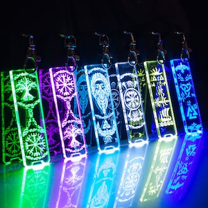 May include: Seven colourful acrylic keychains featuring various Norse symbols and designs. The keychains are illuminated from behind, creating a glowing effect. The symbols include a tree of life, a compass, a runic knot, and a ship.