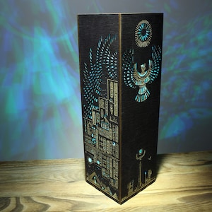 May include: A wooden lamp with a black finish and intricate laser cut designs. The design features an Egyptian-inspired motif with a winged creature and hieroglyphics. The lamp is illuminated with a blue light.