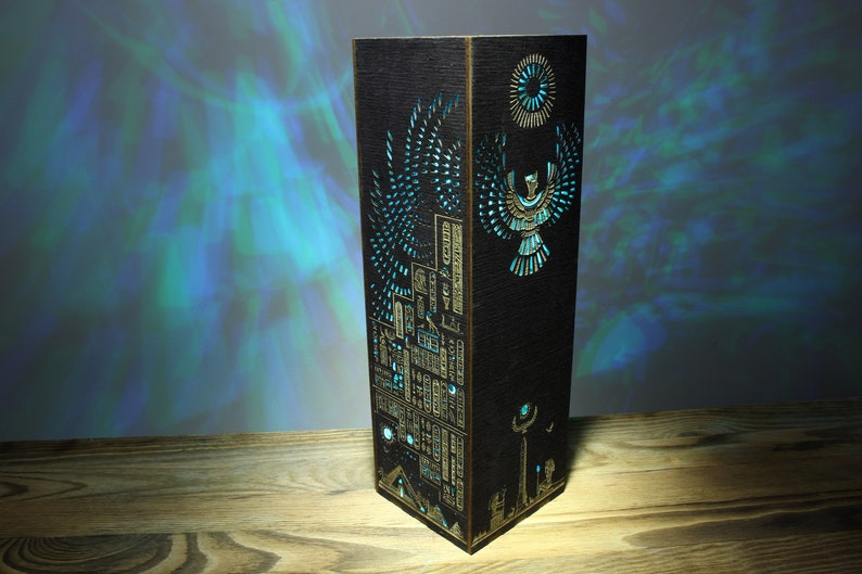 May include: A wooden lamp with a black finish and intricate laser cut designs. The design features an Egyptian-inspired motif with a winged creature and hieroglyphics. The lamp is illuminated with a blue light.