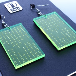 K&ouml;nnte beinhalten: Ein Paar rechteckige, neongr&uuml;ne Ohrringe mit silbernen Haken. Jeder Ohrring hat ein Muster aus wei&szlig;em Bin&auml;rcode. Die Ohrringe werden auf einer dunklen Oberfl&auml;che mit einem kleinen Logo in der oberen linken Ecke pr&auml;sentiert.