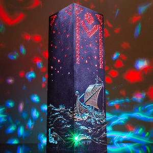 Op de afbeelding: Een hoge, donkere, rechthoekige lamp met uitgesneden ontwerpen van een Vikingschip op golven en hemelse patronen. De lamp straalt een levendige, veelkleurige lichtprojectie uit van rode, blauwe en groene sterren en vormen in de omgeving.
