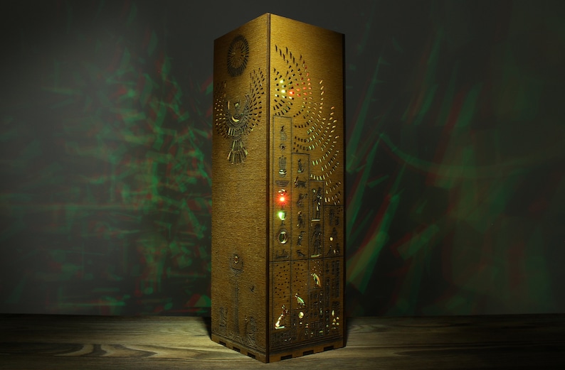 May include: A wooden lamp with intricate carvings of Egyptian hieroglyphs and a winged sun god. The lamp is illuminated from within, casting a warm glow.