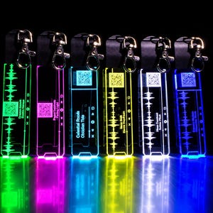 Peut inclure: Six porte-clés rectangulaires en acrylique avec fermoirs mousquetons en métal, illuminés en vert, rose, cyan, jaune, blanc et bleu. Chaque porte-clé présente un graphique d'onde sonore, un code QR et du texte imprimé.