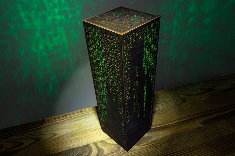 May include: A black cube-shaped lamp with a green light shining through the sides. The sides of the cube are covered in a pattern of letters and numbers.