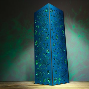 May include: A blue wooden lamp with a cutout design of sea turtles and seaweed. The lamp is illuminated with a green light, casting shadows on the wall behind it.
