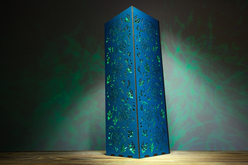 May include: A blue wooden lamp with a cutout design of sea turtles and seaweed. The lamp is illuminated with a green light, casting shadows on the wall behind it.