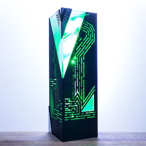 Op de afbeelding: Een hoog, zwart decoratief object met een geometrisch ontwerp en heldergroene LED-verlichting. Het ontwerp lijkt op patronen van printplaten. Het object is rechthoekig met een driehoekige bovenkant en staat tegen een lichte achtergrond.