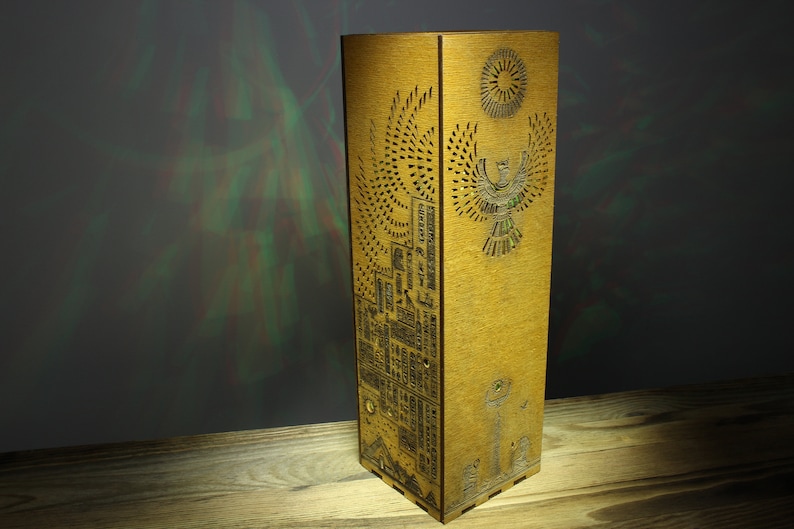 May include: A wooden lamp with intricate carvings of Egyptian hieroglyphs and a winged sun god. The lamp is a light brown color and has a rectangular shape.