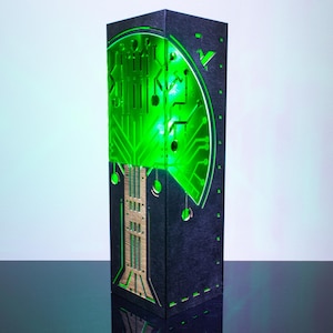 Peut inclure: Une lampe rectangulaire haute avec un extérieur sombre et texturé présentant des motifs de circuits imprimés découpés au laser. Le panneau avant est éclairé de l'intérieur par une lumière verte vive, soulignant les motifs électroniques.