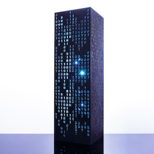 May include: A tall, rectangular black pillar featuring a vertical display of glowing blue binary code consisting of ones and zeros. The surface of the object has a dark, textured finish, and the illuminated digital pattern creates a bright contrast.