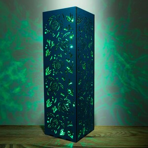 May include: A blue wooden lamp with a cutout design of sea turtles and seaweed. The lamp is lit from within, casting a green glow.