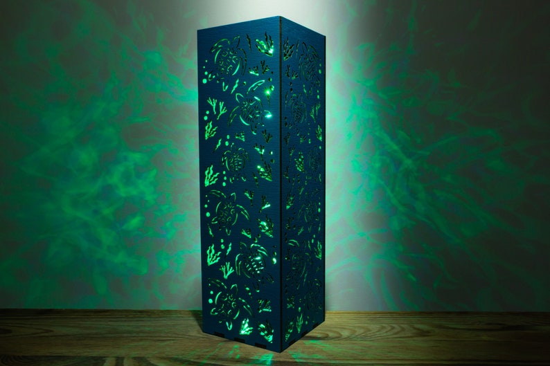 May include: A blue wooden lamp with a cutout design of sea turtles and seaweed. The lamp is lit from within, casting a green glow.