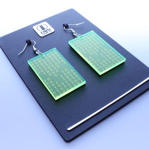 K&ouml;nnte beinhalten: Ein Paar rechteckige, neongr&uuml;ne Ohrringe mit einem Bin&auml;rcode-Muster. Die Ohrringe h&auml;ngen an silbernen Haken und werden auf einer dunkelgrauen Oberfl&auml;che pr&auml;sentiert. Die Ohrringe haben ein modernes, technikbasiertes Design.