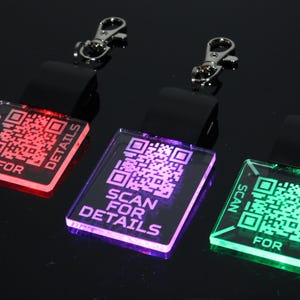 Puede incluir: Tres llaveros iluminados con códigos QR. Cada llavero tiene un color diferente: rojo, morado y verde. El texto "SCAN FOR DETAILS" se muestra en cada llavero. Cada llavero tiene una correa negra y un cierre plateado.
