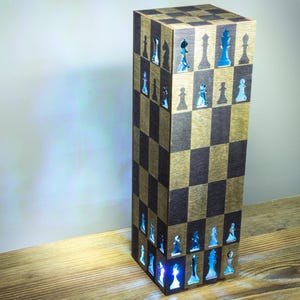 Könnte beinhalten: Eine hohe, rechteckige Lampe mit einem Schachbrettmuster in hellen und dunklen Holztönen auf der Außenseite. Die Oberfläche ist mit Ausstanzungen in Form von Schachfiguren versehen, die ein hellblaues Licht aus dem Inneren abgeben.