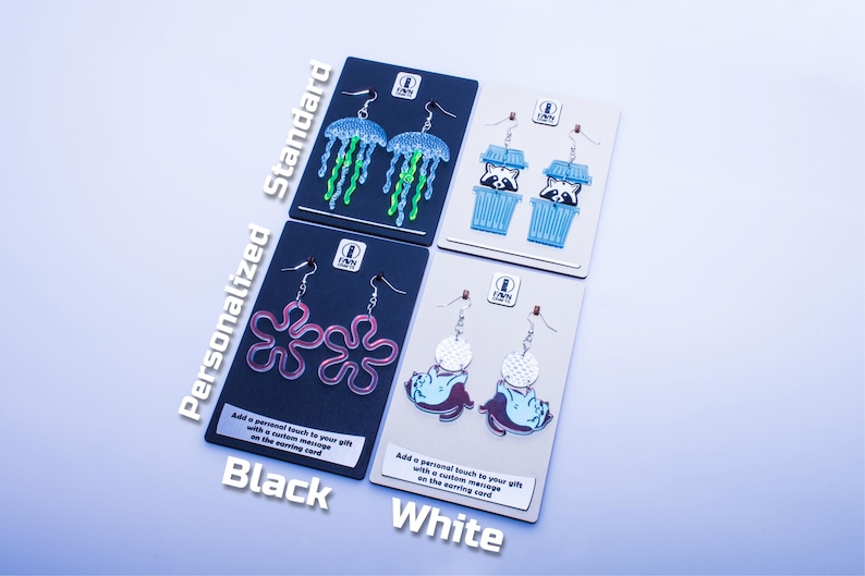 K&ouml;nnte beinhalten: Vier Paar Ohrringe auf Karten pr&auml;sentiert. Die Designs umfassen Quallen, Waschb&auml;ren in Bechern, rosa Blumenformen und ein Wesen mit einem gepunkteten Kreis. Die Karten sind schwarz und wei&szlig;, mit dem Text "Personalized Standard Black White".