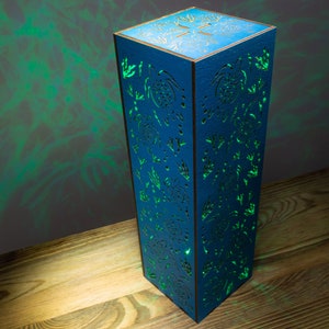 May include: A blue wooden lamp with a cutout design of trees and leaves. The lamp is illuminated with a green light, casting shadows on the wooden surface.
