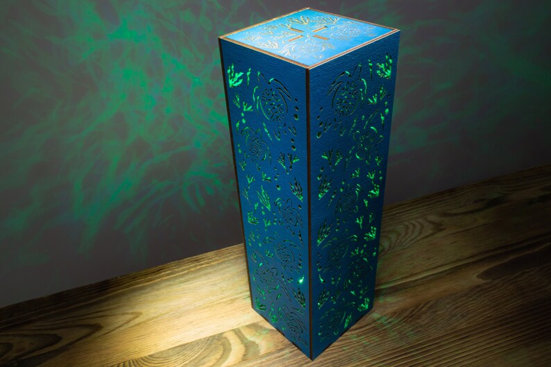 May include: A blue wooden lamp with a cutout design of trees and leaves. The lamp is illuminated with a green light, casting shadows on the wooden surface.